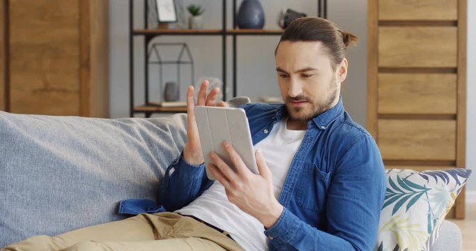 Portrait shot of the young handsome caucasian man lying on the sofa with pillows, scrolling and reading from the tablet device screen. At home. Indoors