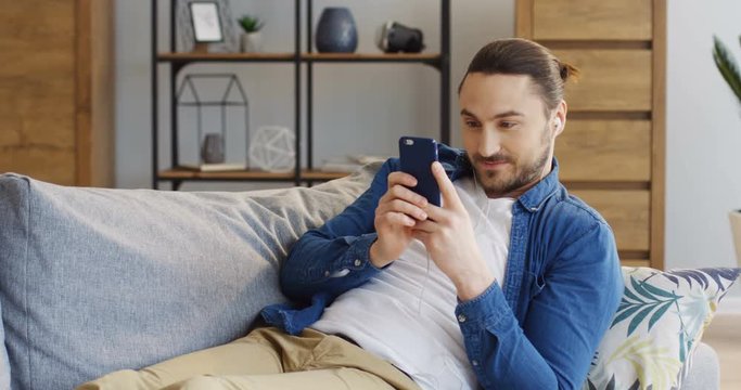 Young attractive man in the headphones resting on the couch with pillows and laughing while watching something on the smartphone screen vertically. At home. Indoor