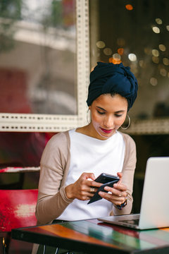 Portrait of a slim, young Muslim Malay woman sitting and working at a cafe during the day. She is wearing a turban and is fashionably dressed.