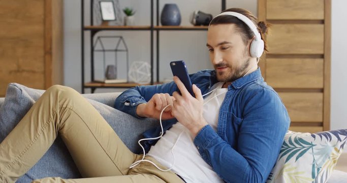 Caucasian young man in the big white headphones and casual style looking at the smartphone in his hands. Cozy room. Indoors