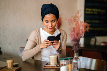 Portrait of a young Muslim Malay woman sitting in a cafe and using her smartphone as she enjoys her morning coffee. She is elegantly and fashionably dressed and is wearing a turban hijab. 
