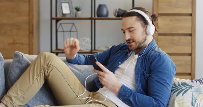 Handsome young man in the big white headphones and casual style singing and looking at the smartphone screen in his hands. Cozy living room. Inside