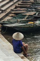 Back of woman wearing purple shirt, conical hat carry wash her feet in the river with empty rowing boats in the background at Trang An Grottoes in Ninh Binh, Vietnam.