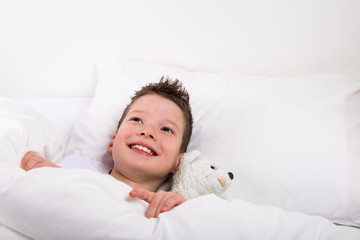 smiling boy resting in a white bed together with toy  a white teddy bear