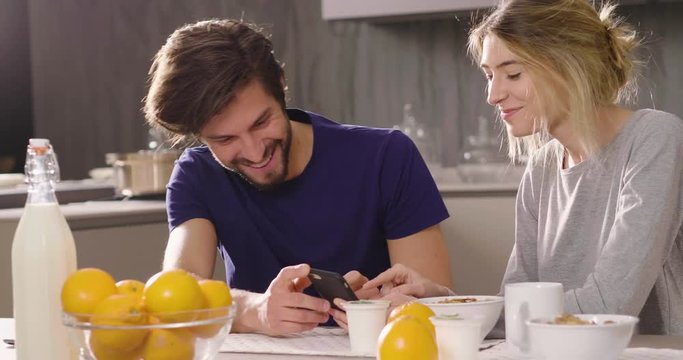 A couple in the kitchen looks at the telephone with the souvenir photos of their holidays or of the past times while having breakfast and smiling happily. Concept of: family, technology, memories.