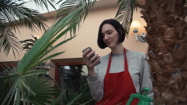 Young woman gardener in red apron using smartphone in home garden