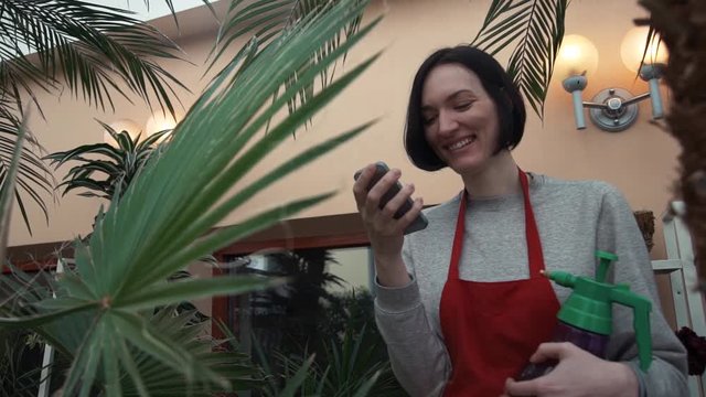 Young smiley girl in red apron laughing using smartphone in greenhouse