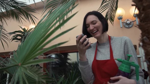 Young florist girl in red apron laughing using smartphone in greenhouse