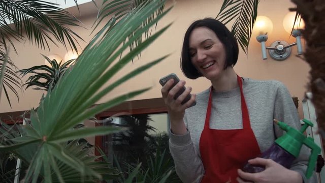 Young florist girl in red apron laughing using smartphone in house garden