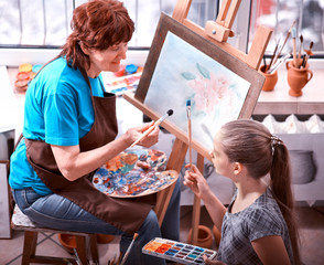 Fototapeta premium Artist painting easel in studio. Authentic grandmother and kids girl paints with palette watercolor paints palette and brush morning sunlight. Courses on drawing for children and adults.