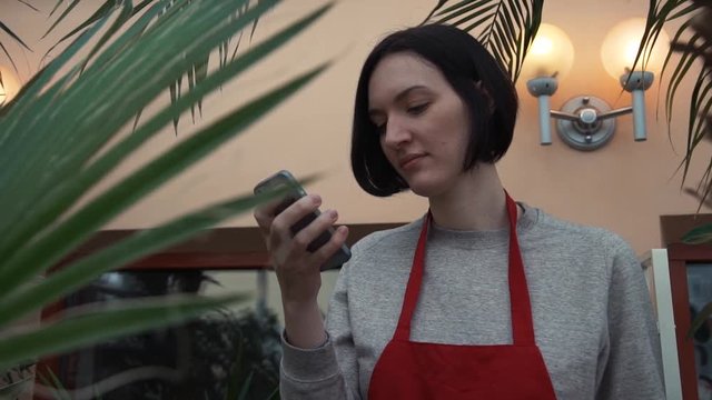 Portrait of young woman in red apron using smartphone in greenhouse.