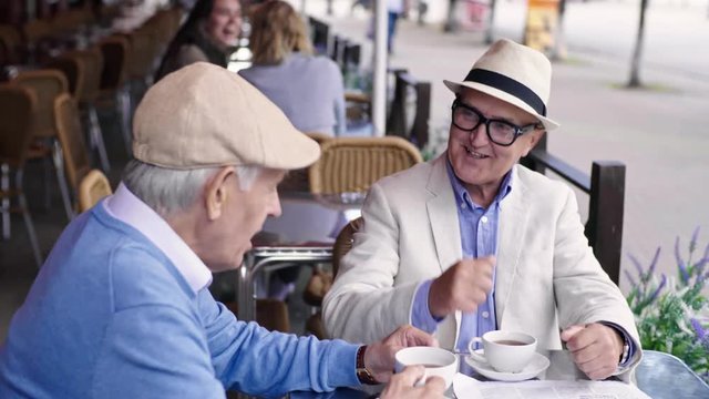 Medium shot of two senior men in hats sitting next to each other at table in street cafe and talking over cup of tea