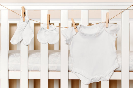 Set of baby clothes for newborn in white color. Baby socks, mittens and bodysuit are hanging on a clothline with infant bed on background. Newborn concept background.