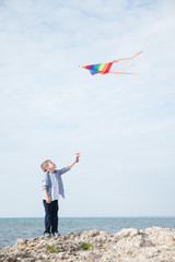 handsome little kid holding flying colorful kite standing on rock near open sea