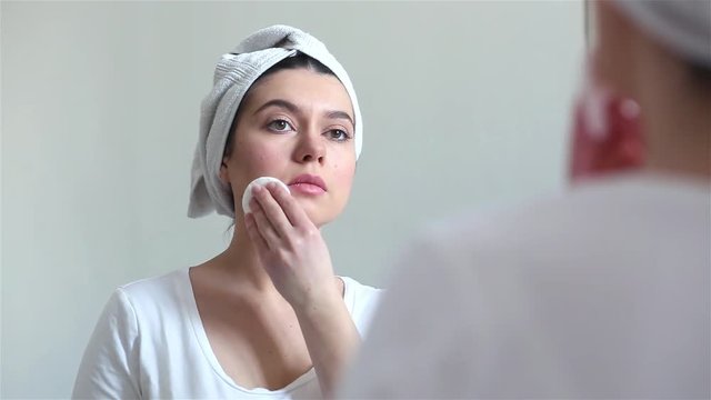 Young brunette woman with towel on hair removing makeup from her face, skin care concept.
