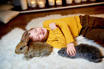 Child playing with grey rabbit. Young boy lying relaxed with his eyes shut, two grey rabbits sitting next to him. Easter celebration. Children and animals. Kids take care of pets