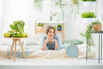 Portrait of a young and beautiful woman lying in the bright and cozy bedroom with fresh green food and plants at home