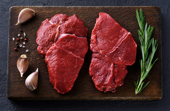 Fresh raw beef steaks, pepper, garlic, salt and rosemary on a dark wooden background.