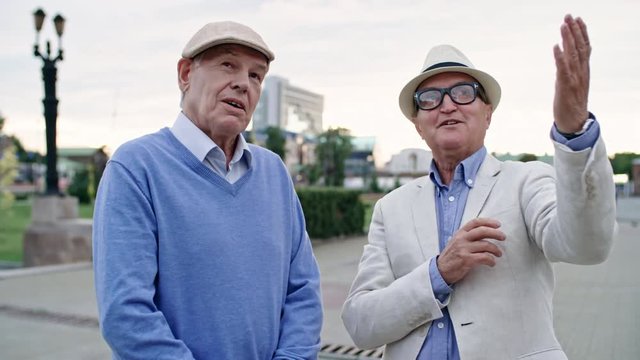 Two retired male friends in hats standing in the street and talking about surroundings, medium shot