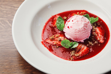 beautiful and appetizing horizontal close-up image of round ice cream scoop pink in plate supplemented with red sauce, pieces of strawberries and mint leaves, sprinkled with dessert crumbs.