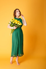 pensive woman in green dress with bouquet of yellow tulips isolated on orange