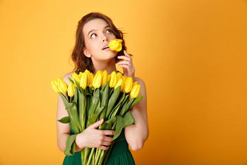 portrait of pensive woman with tulip in mouth and bouquet of yellow tulips isolated on orange