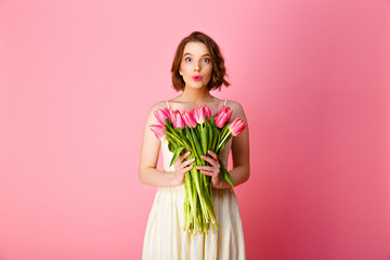 portrait of grimace woman in white dress holding bouquet of pink tulips isolated on pink