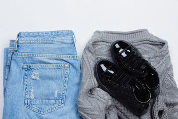 Woman Trendy Fashion Clothes. Blue jeans, knitted gray sweater, patent leather shoes boots on white background, flat lay, top view, copy space