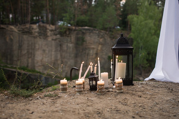 A lot of white burning candles on candlesticks and in lanterns in forest near the wedding arch with white material
