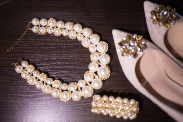 Beautiful set of women's wedding accessories. Pearl necklace in focus, beige shoes with shiny stones on them, and a bracelet on the wooden background. Accessories