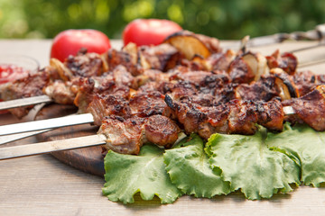 Grilled pork on skewers roasted on the grill and lettuce leaves on wooden chopping board with tomato sauce in a bowl and tomatoes on the background