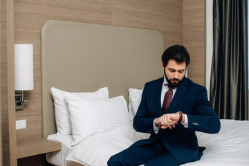 handsome businessman sitting on bed at hotel room and looking at watch