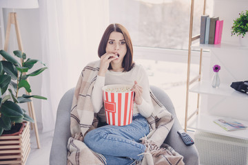 Naklejka premium Portrait of pretty, charming, stylish, exciting girl with modern hairstyle putting pop corn in her open mouth watching spectacular film, covered with plaid, wearing denim outfit, jeans and pullover