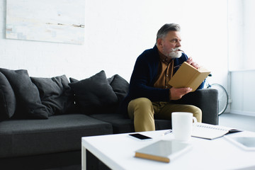 pensive bearded senior man holding book and looking away at home