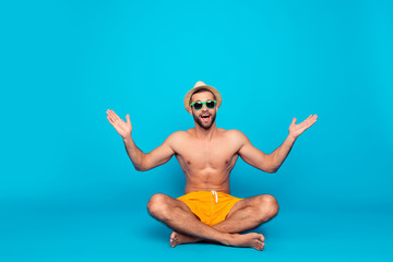 Cheerful, funny, attractive, ladies' man with naked torso in yellow shorts sitting with crossed legs over blue background, enjoying, admire weather, nature, holidays with raised hands and open mouth