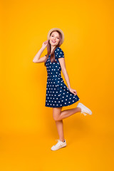 Full-length, fullbody portrait of nice, cheerful, positive, cute girl with raised leg, holding hands on hat and bottom of polka-dot dress, posing over yellow background