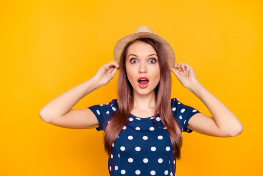 Portrait of charming, pretty, nice, impressed, shocked girl in polka-dot t-shirt holding hat with two hands, having wide open mouth and eyes, isolated on yellow background