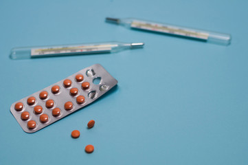 Two glass mercury thermometers and blister pack with brown round pills or drugs and scattered pills on blue background with copy space