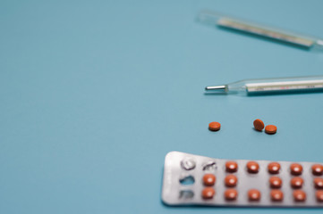 Two glass mercury thermometers and blister pack with brown round pills or drugs and scattered pills on blue background with copy space