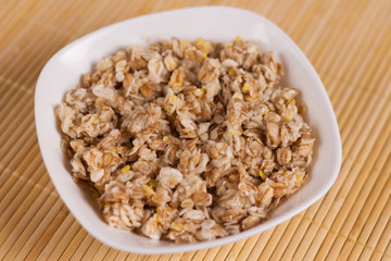 Cooked yummy warm oatmeal in white ceramic bowl on clean bamboo rug