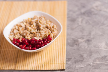 Cooked yummy warm oatmeal and heap of ripe fresh pomegranate seeds in white ceramic bowl on clean bamboo rug on worn gray scratched concrete with copy space