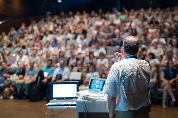Speaker giving a talk on corporate business conference. Unrecognizable people in audience at...