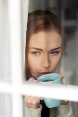 Beautiful caucasian woman drinking hot coffee or tea and looking through window.