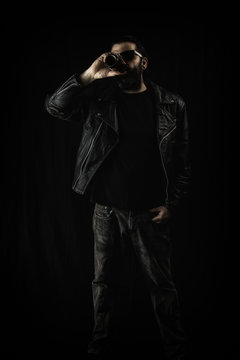 A man wearing a black leather jacket and sunglasses, drinking from a bottle of beer.