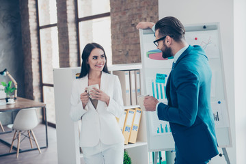 Handsome man in glasses with bristle making advances to his charming colleague, lean with hand on flip-chart, white board, lovely couple speaking during freetime holding mug with tea in hands