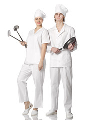 A beautiful young couple dressed in cooks with kitchen tools stand at full length next to each other.
