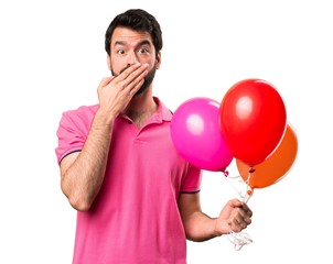 Handsome young man holding balloons and covering his mouth over isolated white background