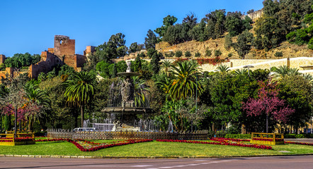Obraz premium Fountain (Fuente de las tres gracias) at the end of Paseo del Parque with the castle (alcazaba) to rear. Malaga city, Spain.