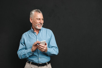 Image of bearded male pensioner 60s with gray hair chatting or browsing internet on mobile phone, isolated over black background
