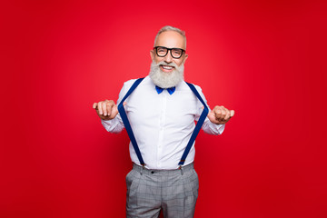 Portrait of elegant, confident, playful macho, old barber, stylist draw off suspenders with thumb fingers, looking at camera with beaming smile, isolated on red background